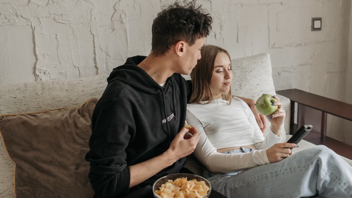 A young couple enjoys snacks while relaxing on a sofa, using a smartphone indoors.